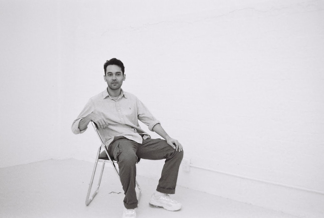 A black and white image of Robbie leaning back in a chair in an empty room. Looking at the camera, holding a slight smile. Robbie wears a button up long sleeve top, with the top button undone and sleeves rolled up. He is wearing dark cargo pants with white Nike sneakers. His hand is placed on his knee.